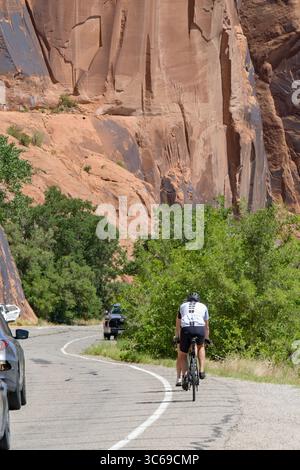 Moab, Utah, USA - 25. Mai 2025: Radfahrer radeln auf der Potash Road unter riesigen Felsen in der Nähe von Moab mit vorbeifahrendem Verkehr Stockfoto