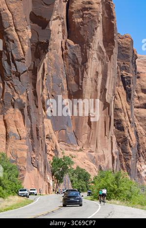 Moab, Utah, USA - 25. Mai 2025: Radfahrer radeln auf der Potash Road unter riesigen Felsen in der Nähe von Moab mit vorbeiziehendem Verkehr Stockfoto