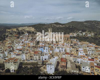 Blick aus der Vogelperspektive auf alte Steinbauten, die einen Hügel hinunter zu einer zentralen Kirche und einem Turm stürzen, im Kontrast zu den umliegenden grünen Hügeln, Cervera del Maestre, Valencianische Gemeinde, Spanien. Stockfoto