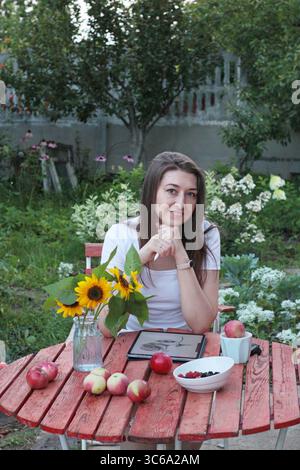 Junge Künstlerin, die an einem roten Holztisch im Garten mit Tablette, Äpfeln, Beeren, Sonnenblumen und Tablette auf weißem Hintergrund sitzt. Konzept des Sommers Stockfoto