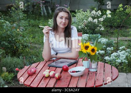 Junge Künstlerin sitzt an einem roten Holztisch im Garten mit Äpfeln, Beeren, Sonnenblumen und Tablette auf weißem Hintergrund. Konzept der Sommerzeichnung, Stockfoto