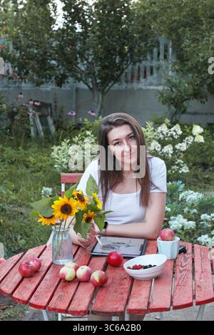 Junge Künstlerin, die an einem roten Holztisch im Garten mit Tablette, Äpfeln, Beeren, Sonnenblumen und Tablette auf weißem Hintergrund sitzt. Konzept des Sommers Stockfoto