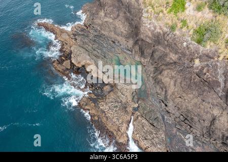 Aus der Vogelperspektive auf die zerklüftete Küste, wo die dunklen, strukturierten Felsen auf das türkisfarbene Wasser treffen, eine versteckte Bucht, die von oben sichtbar wird, Whangarei, Northland Region, Neuseeland. Stockfoto