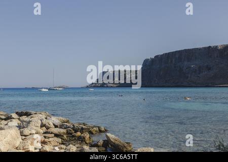 Der Blick auf das türkisfarbene Wasser ragt sanft an eine felsige Küste, im Kontrast zu den imposanten Klippen und weit entfernten Segelbooten, Lindos, Rhodos, Griechenland. Stockfoto