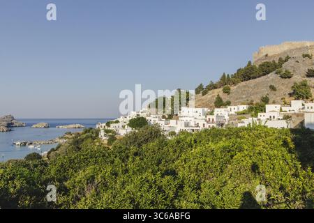 Blick auf helle weiße Gebäude, die den Hügel hinunter zum türkisfarbenen Meer kaskadieren, gekrönt von antiken Steinruinen in Lindos, Rhodos, Griechenland. Stockfoto