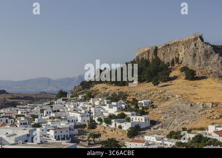 Blick auf weiß getünchte Gebäude, eingebettet unter den zerklüfteten, sonnenverwöhnten Klippen, gekrönt von antiken Ruinen vor einem klaren blauen Himmel, Lindos, Rhodos, Griechenland. Stockfoto