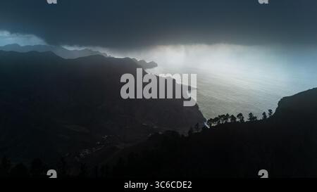 Aus der Vogelperspektive auf den Regen, der über die zerklüftete Küste zieht und ein dramatisches Licht über die dunklen Klippen und den turbulenten Ozean wirft, Agaete, die Kanarischen Inseln, Spanien. Stockfoto