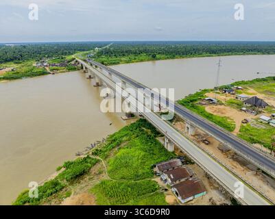 Aus der Vogelperspektive auf eine riesige Brücke, die sich über das braune Wasser erstreckt, flankiert von üppigen grünen Ufern und weit entfernten Bäumen, Yenegoa, Bayelsa, Nigeria. Stockfoto