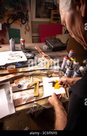 Ein älterer Künstler, der in einem bunten, urbanen Studio mit Sprühfarbe an goldenen Zahlenschablonen arbeitet. Ideal für Kreativität, Handwerkskunst, ... Stockfoto