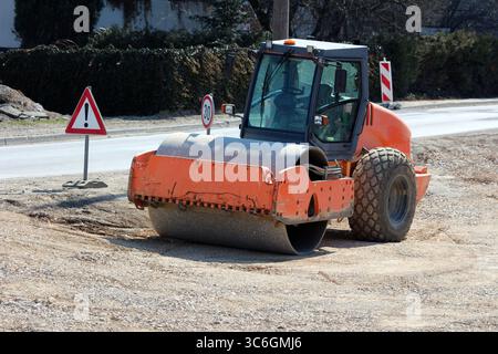 Eine orangene Eintrommel-Vibrationswalze verdichtet Kies und Boden auf einer aktiven Baustelle, begleitet von Warnschildern und einer Geschwindigkeit von 30 km Stockfoto