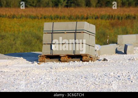 Ein Bündel rechteckiger, mit schwarzen Bändern gebundener Betonfertigteile befindet sich auf einer Holzpalette, die von Kies umgeben ist, an einer Straßenbaustelle Stockfoto
