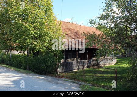 Ein rustikales Holzhaus mit rotem Ziegeldach liegt im Schatten von Bäumen und umgeben von einem Pflaumenzaun, das traditionelle ländliche Architektur vereint Stockfoto