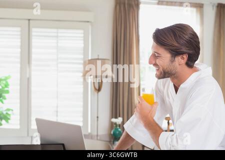 Männlicher Erwachsener, der sich im Wohnzimmer auf den Tisch lehnt und Orangensaft hält, während er im Laptop surft, Kopierraum. Gemütlich, lässig, minimalistisch, Entspannung, Home Office, Stockfoto