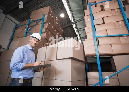 Männlicher Mann mittleren Alters mit Schutzhelm im Lager, der die Zwischenablage zwischen den Kästen und blauen Regalen inspiziert. Industrie, Logistik, Organisation, Sicherheit, Invento Stockfoto