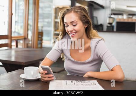 Frau, die an einem Holztisch im Café sitzt, trägt eine graue Oberseite und lächelt das Smartphone neben der Kaffeetasse an. Lässig, Lifestyle, rustikal, modern, sozial, l Stockfoto