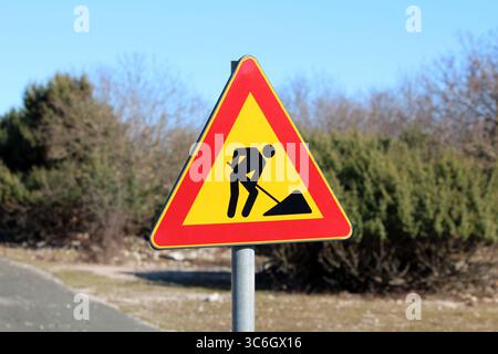 Ein helles dreieckiges Straßenschild mit einem schwarzen Bauarbeitersymbol warnt die Fahrer vor bevorstehenden Bauarbeiten und hebt sich von grünen Sträuchern ab Stockfoto