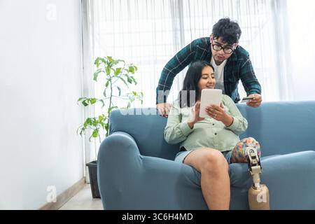 Schwangere Frau mit einer Beinprothese, die ein Tablet benutzt und Online-Einkäufe tätigt, während ihr Mann eine Kreditkarte hält Stockfoto