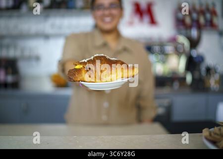 Eine Person hält ein Croissant auf einem Teller in Richtung der Kamera in einem Café, mit sanft verschwommenem Hintergrund, Cremona, Italien 25. Juli 2025 Stockfoto