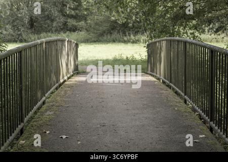 Schmale Metallbrücke, die über ein kleines, trockenes Flussbett in einem ruhigen Sommerpark spannt, lädt zum Erkunden und friedlichen Spaziergängen ein Stockfoto