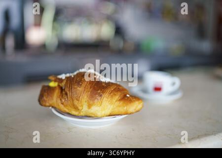 Nahaufnahme eines Croissants auf einem weißen Teller, platziert auf einem Café-Theke mit sanft fokussiertem Hintergrund, Cremona, Italien 25. Juli 2025 Stockfoto