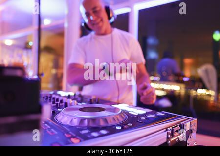 Verschwommener DJ mit Kopfhörern an einem Mischpult mit Plattenspieler und Bedienelementen macht Musik bei einer Veranstaltung in stimmungsvoller Partyatmosphäre mit farbenfrohem Licht Stockfoto