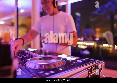 Verschwommener DJ mit Kopfhörern an einem Mischpult mit Plattenspieler und Bedienelementen macht Musik bei einer Veranstaltung in stimmungsvoller Partyatmosphäre mit farbenfrohem Licht Stockfoto