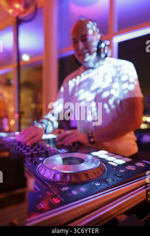 Verschwommener DJ mit Kopfhörern an einem Mischpult mit Plattenspieler und Bedienelementen macht Musik bei einer Veranstaltung in stimmungsvoller Partyatmosphäre mit farbenfrohem Licht Stockfoto
