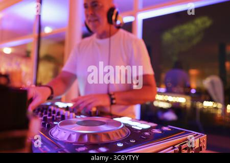 Verschwommener DJ mit Kopfhörern an einem Mischpult mit Plattenspieler und Bedienelementen macht Musik bei einer Veranstaltung in stimmungsvoller Partyatmosphäre mit farbenfrohem Licht Stockfoto
