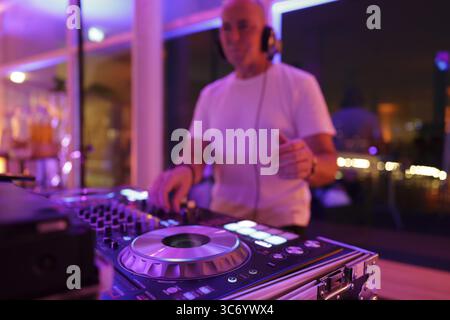 Verschwommener DJ mit Kopfhörern an einem Mischpult mit Plattenspieler und Bedienelementen macht Musik bei einer Veranstaltung in stimmungsvoller Partyatmosphäre mit farbenfrohem Licht Stockfoto