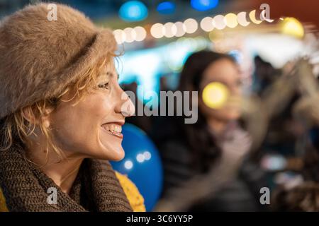 Profil einer fröhlichen, erwachsenen Frau, die eine Pelzmütze und einen Schal trägt und unter bunten Bokeh-Lichtern auf einem Wintermarkt lacht. Stockfoto