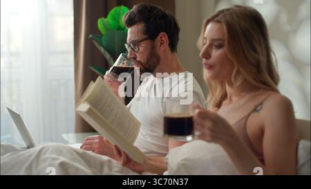 Familie ruhig kaukasisches Paar trinkt Kaffee im Bett Schlafzimmer Morgenbeziehung Frau las Buch Literatur Paar entspannende Bettwäsche Mann mit Laptop Stockfoto