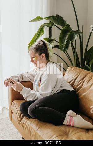 Frau sitzt auf der Couch mit Augenmasken, während sie das Handy benutzt Stockfoto