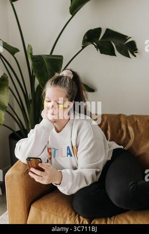 Frau sitzt auf der Couch mit Augenmasken, während sie das Handy benutzt Stockfoto