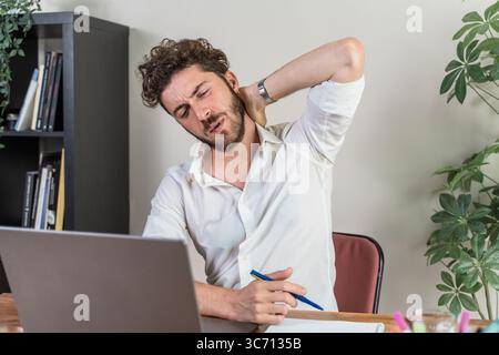 Überarbeiteter junger Geschäftsmann, der sich müde fühlt und unter steifen Nacken- und Rückenschmerzen leidet, aufgrund schlechter Körperhaltung und langer Arbeitsstunden am Computer in Stockfoto