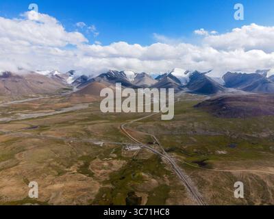 Ein weites Panorama auf eine abgelegene Berglandschaft mit unbefestigten Straßen, verstreuten Feuchtgebieten und einer Reihe von schneebedeckten Gipfeln Stockfoto