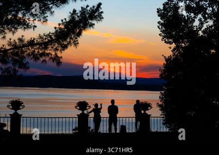 Silhouetten von Menschen, die an einem Geländer stehen und einen lebhaften Sonnenuntergang über dem Bodensee in der Schweiz bewundern. Der strahlende Himmel spiegelt sich auf dem ruhigen Wasser Stockfoto