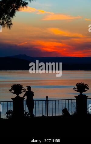 Silhouette einer Frau, die sich auf einem Geländer stützt und einen lebhaften Sonnenuntergang über dem Bodensee in der Schweiz genießt. Der dramatische orange-rosa Himmel spiegelt sich wider Stockfoto