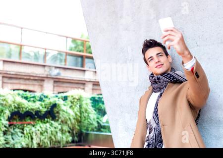 Kaukasischer junger Mann in beigem Mantel und Schal, der ein Selfie mit Smartphone macht, an einer Betonmauer in einer städtischen Umgebung steht, lächelnd und selbstbewusst. Stockfoto