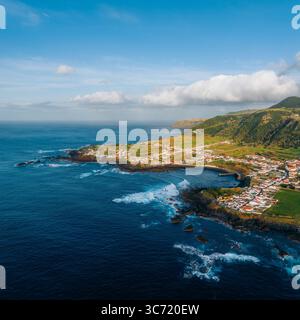Blick aus der Vogelperspektive auf die zerklüftete Küste, wo dunkle vulkanische Felsen auf das turbulente türkisfarbene Meer treffen, im Kontrast zu den grünen Hügeln in Mosteiros, Portugal. Stockfoto