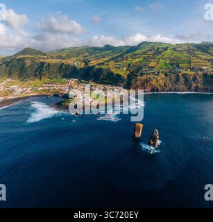 Blick aus der Vogelperspektive auf die dramatische Küste, die in der Nähe von Ilhéus dos Mosteiros auf das weite Meer trifft, mit der Stadt eingebettet in die grünen Hügel, Mosteiros, Portugal. Stockfoto
