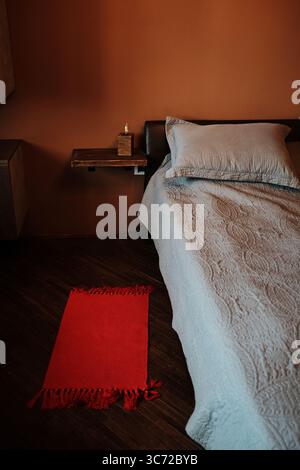 Minimalistische Schlafzimmerecke mit blauer Bettwäsche, orangefarbenen Wänden, hölzernem Nachttisch und hellem rotem Teppich auf dunklem Holzboden. Gemütliche und warme Inneneinrichtung. Stockfoto