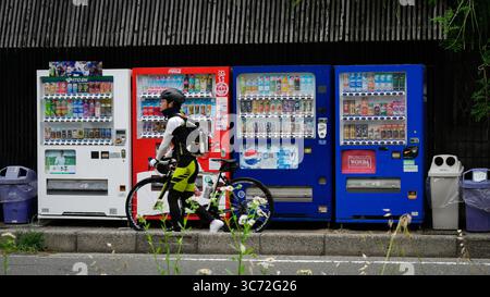Radfahrer vorbei an farbenfrohen japanischen Automaten in Kyoto, Japan. Urbane Lifestyle-Szene mit Getränken, Technologie und Alltagsroutine. Stockfoto