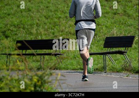 Stuttgart, Deutschland. August 2025. Ein Mann joggt auf einem Asphaltweg in einem Park. Im Hintergrund stehen leere Parkbänke vor einem grünen Damm. Quelle: Markus Lenhardt/dpa/Alamy Live News Stockfoto
