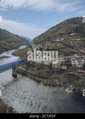Die blaue Brücke aus der Vogelperspektive überspannt den strukturierten Fluss und verbindet das malerische Dorf inmitten der grünen Hügel OS Peares, Ourense, Spanien. Stockfoto