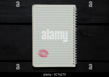 Ein offenes Notizbuch mit Linien auf einer Spirale liegt auf einem Holztisch, unten auf der Seite befindet sich ein klares Küsszeichen mit rotem Lippenstift, Draufsicht Stockfoto