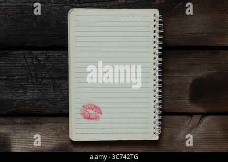 Ein offenes Notizbuch mit Linien auf einer Spirale liegt auf einem Holztisch, unten auf der Seite befindet sich ein klares Küsszeichen mit rotem Lippenstift, Draufsicht Stockfoto