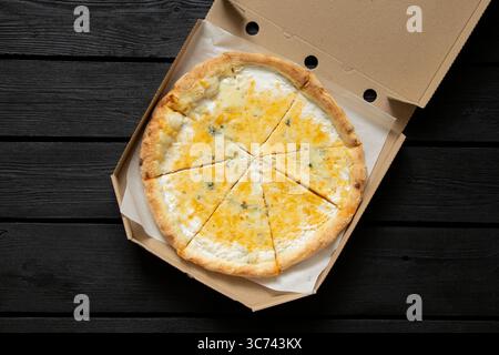 Ganze Käse-Pizza in Stücke geschnitten in einer offenen Box auf einem schwarzen Holztisch Hintergrund, Blick von oben, appetitliches Abendessen Stockfoto