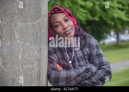 Selbstbewusste, reife Afrikanerin mit überkreuzten Armen, die sich im Park an die Betonsäule lehnt. Schöner 40er-Jahre-Profi mit roten afrikanischen Zöpfen, die c Stockfoto