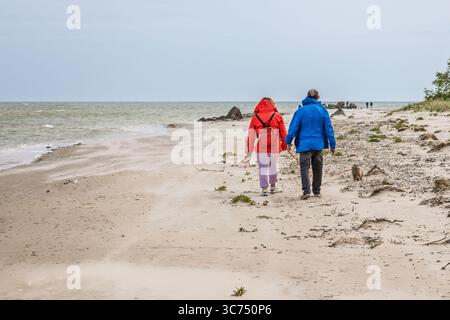 Seniorenpaar in bunten Jacken, die Hand in Hand an einem ruhigen Ostseesandstrand entlang gehen Stockfoto