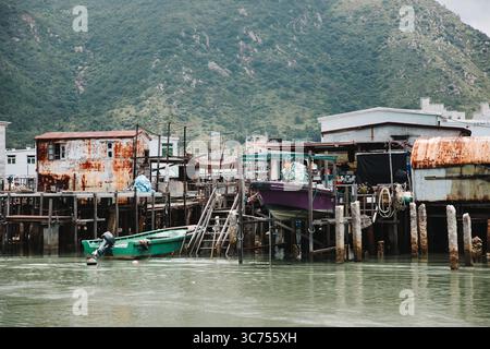 Stelzenhäuser und Fischerboote entlang eines traditionellen Uferdorfes in Hongkong, mit ruhiger Meer- und Bergkulisse. Stockfoto